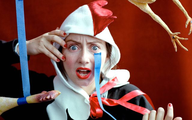 A light-skinned woman wearing a rooster head hood stares in shock at the viewer.