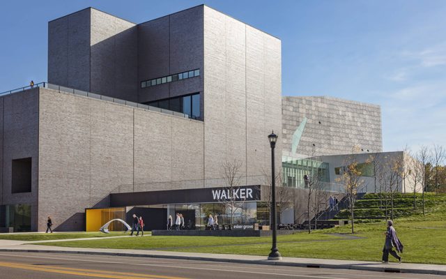 A person walking on a sidewalk in front of the entrance of a gray modern-shaped building, the Walker Art Center.