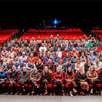 A large theatre with red seats and black stair railings, with mostly the middle section filled with smiling adults posing for a group photo