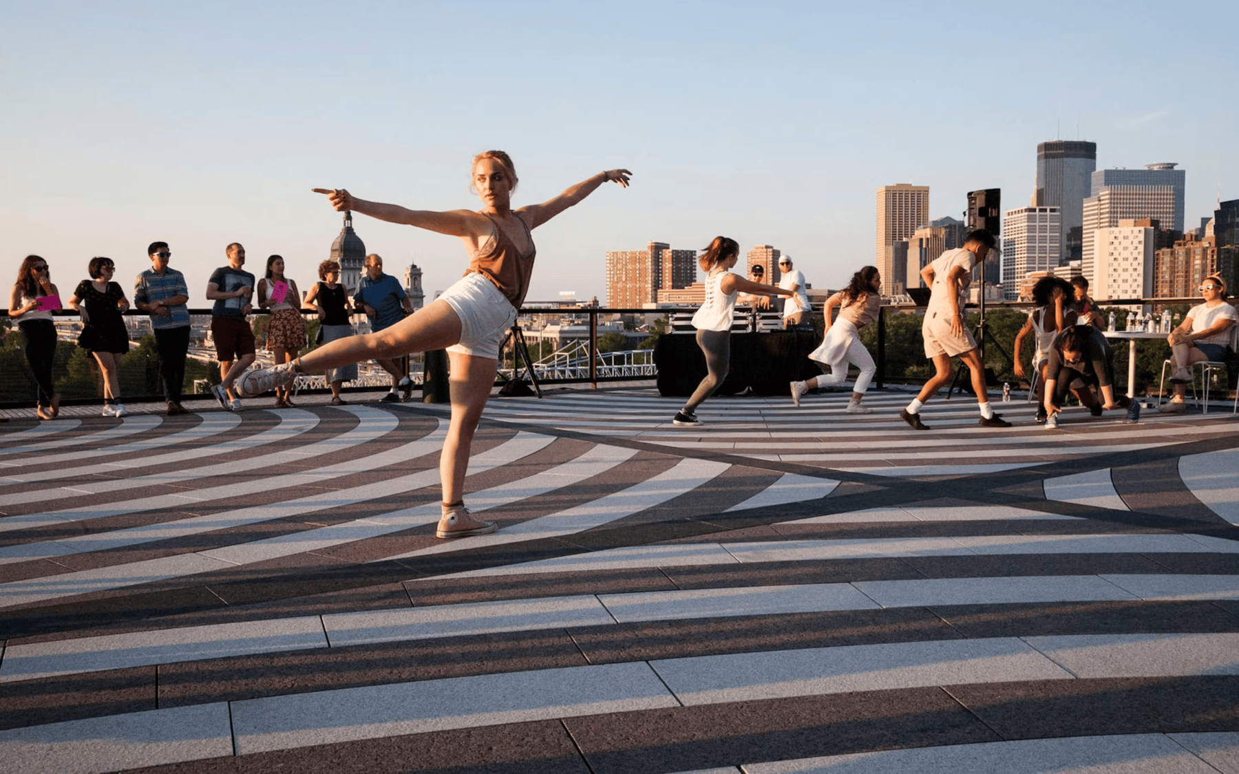 Dancer posed in an arabesque, performing with other dancers in front of a small audience on a striped-designed rooftop during the day.