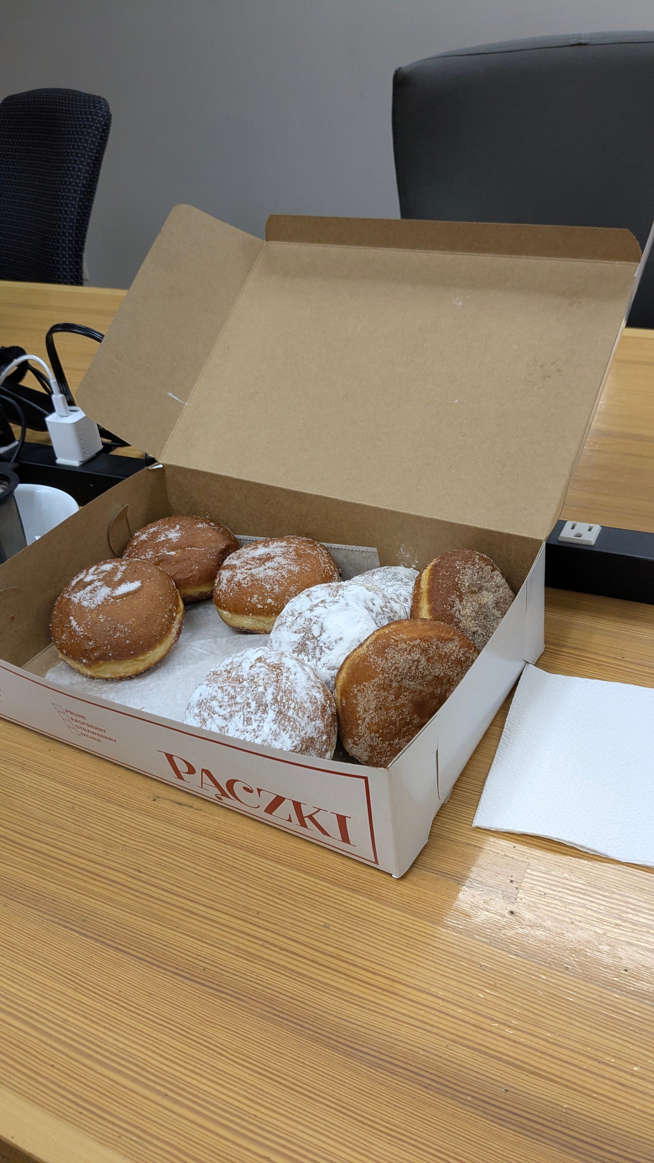 An open box of donuts on a wood conference room table next to plugs and office chairs
