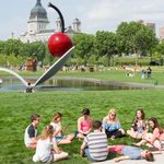 Groups of people enjoying a park with a large outdoor sculpture of a cherry atop a spoon, floating over a small man-made lake, surrounded by grass, trees, and buildings