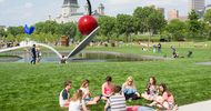 Groups of people enjoying a park with a large outdoor sculpture of a cherry atop a spoon, floating over a small man-made lake, surrounded by grass, trees, and buildings