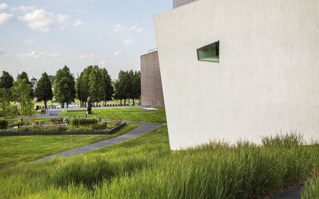 A large grey building next to a smaller white building with a rhombus shaped window next to grass and trees on a sunny day