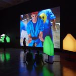 People standing and sitting in a dark room in the middle of light structures and a film installation