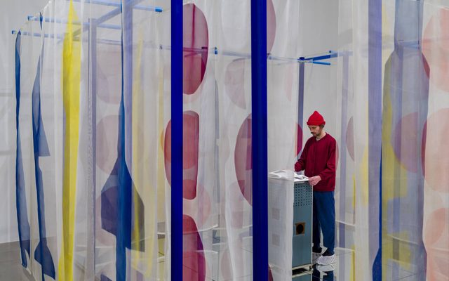 An adult flipping though an art book displayed within an exhibit with blue, yellow, and red shapes on white sheer fabric hanging throughout