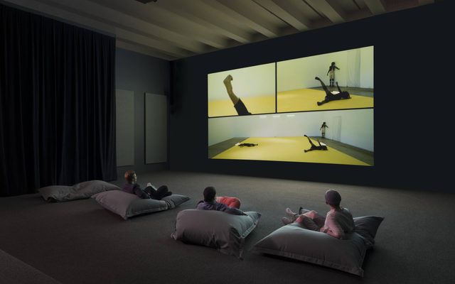 Three people lying on large pillows on the ground, watching a wall-sized screen of another person on a yellow mat doing movements
