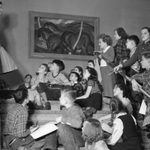 A black and white photo of a classroom of children sitting on the steps of Walker Art Center, listening to their teacher speak.