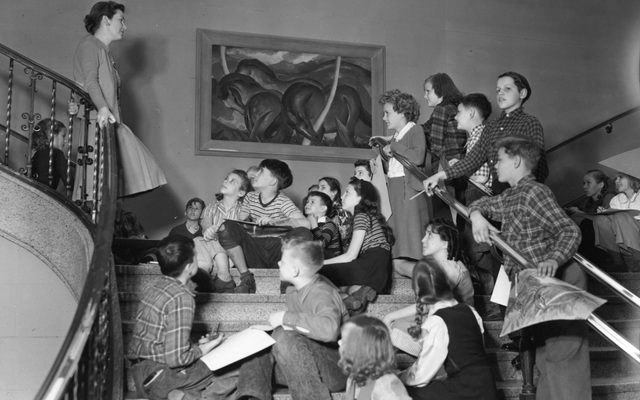 A black and white photo of a classroom of children sitting on the steps of Walker Art Center, listening to their teacher speak.