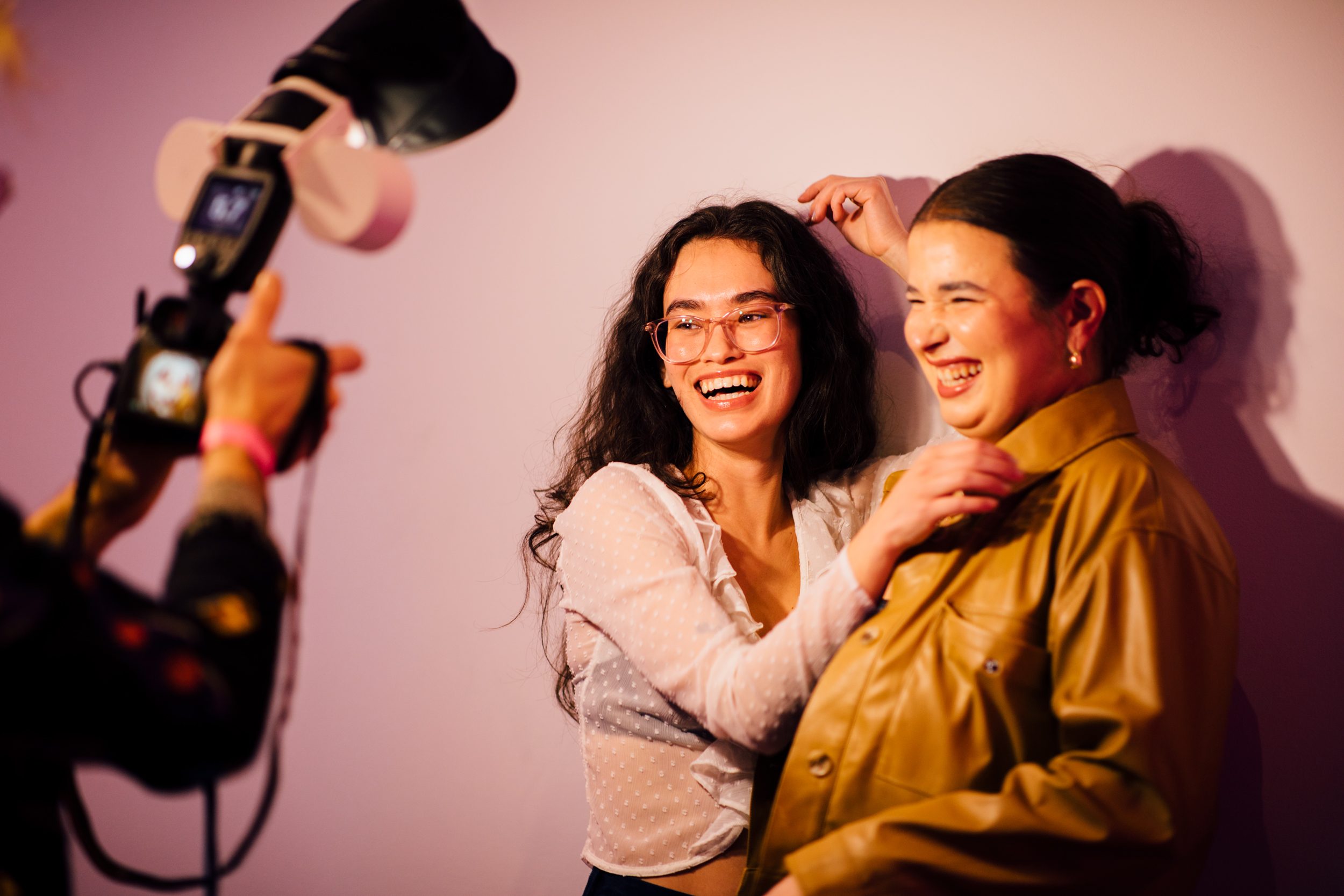 Two people pose against a wall for a camera, which is in view on the left side of the photo. The person on the left smiles at the camera, mid embrace of the person of the left.