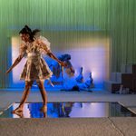 A dancer in a elaborate light colored dress posed standing on top of a mirrored rectangle floor, in front of a blue-lit background with two performers posed on the floor and a chair