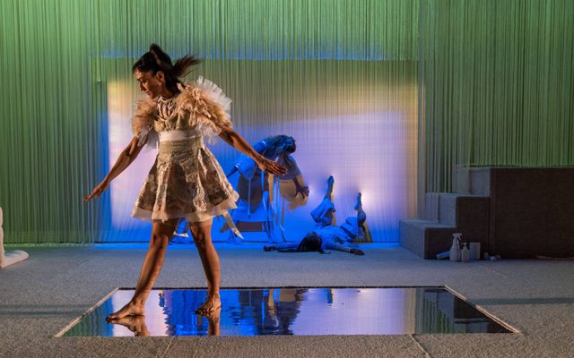 A dancer in a elaborate light colored dress posed standing on top of a mirrored rectangle floor, in front of a blue-lit background with two performers posed on the floor and a chair