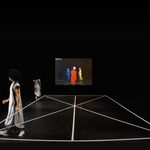 01 Triadic Ballet A dancer in white walks along the edge of a dance floor, her body doubled in the space. A projection along the back wall shows three figures in primary colors.