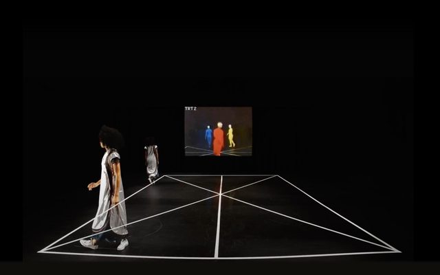 A dancer in white walks along the edge of a dance floor, her body doubled in the space. A projection along the back wall shows three figures in primary colors.