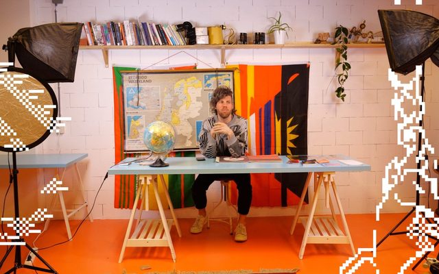 An adult sitting at a table with a globe and colorful backdrop, with lighting and photography equipment around them
