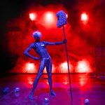 A live artist on stage holding a stick with a parliament-style wig on top, with small silver skulls on the ground in front of smoke and red lights