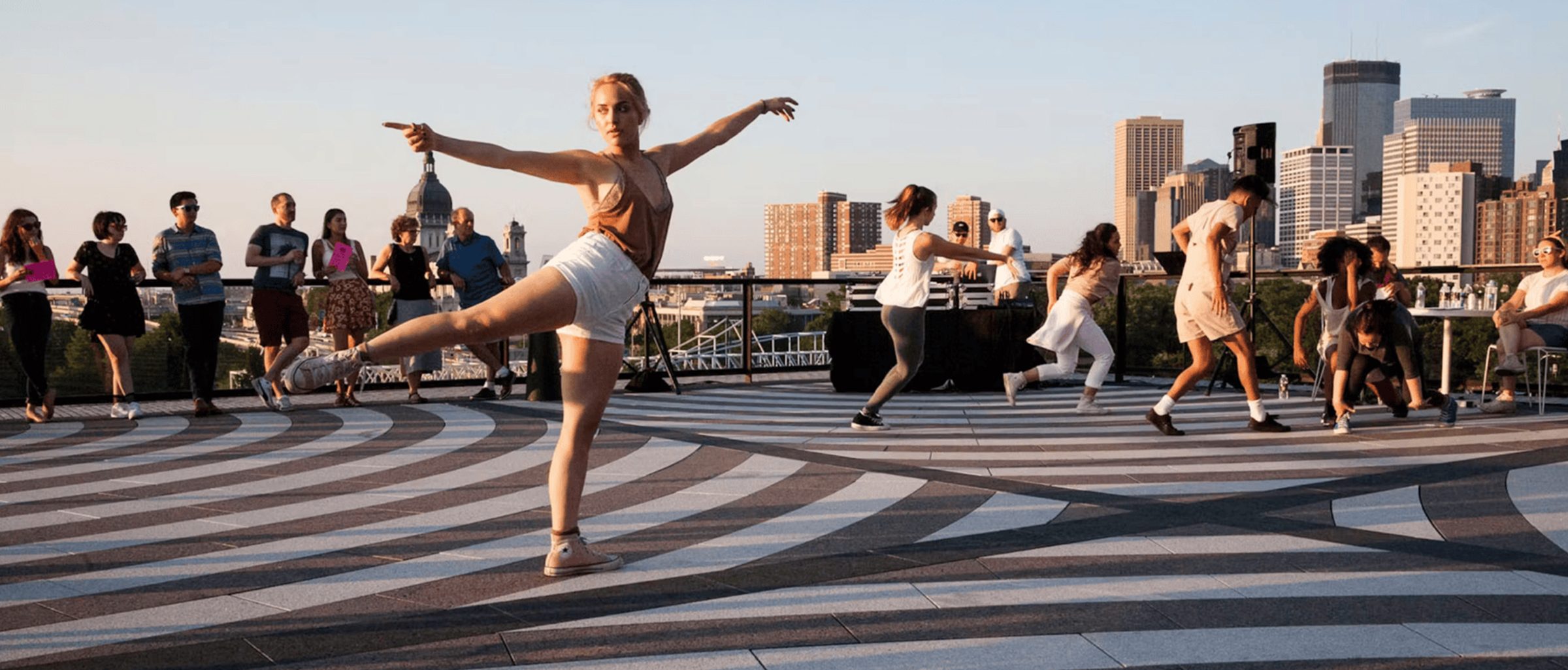 Dancer posed in an arabesque, performing with other dancers in front of a small audience on a striped-designed rooftop during the day.
