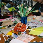 A cluttered table with papers, glue, flowers, and people crafting and cutting out magazine images.