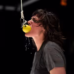 Person with long dark hair bites into a lemon hanging on a hook and rope and lemon juice drips from it