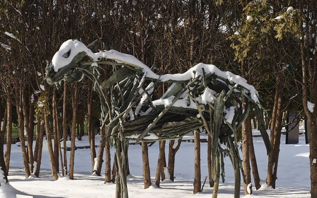 Outdoor sculpture of a large 4 legged animal made of wood branches, covered in snow in a snowy field with trees