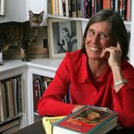 An adult with light skin, wearing a red shirt and silver watch, has medium legth brown hair and smiles at the viewer with a hand to their face. A cat stands on a bookshelf behind.