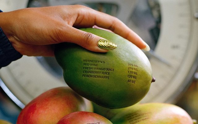 A hand with gold nails holds a mango with cocktail ingredients printed on its skin, above more mangoes.