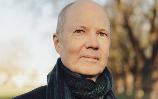 A headshot of an adult with light skin and blue eyes wears a black scarf