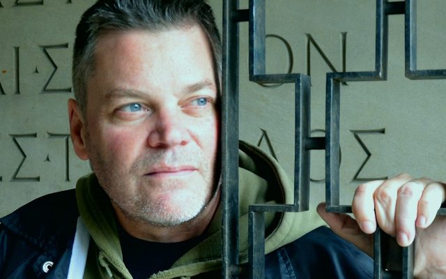 A fair skinned adult with short hair, stubble, and blue eyes holds onto a metal railing and gazing off camera