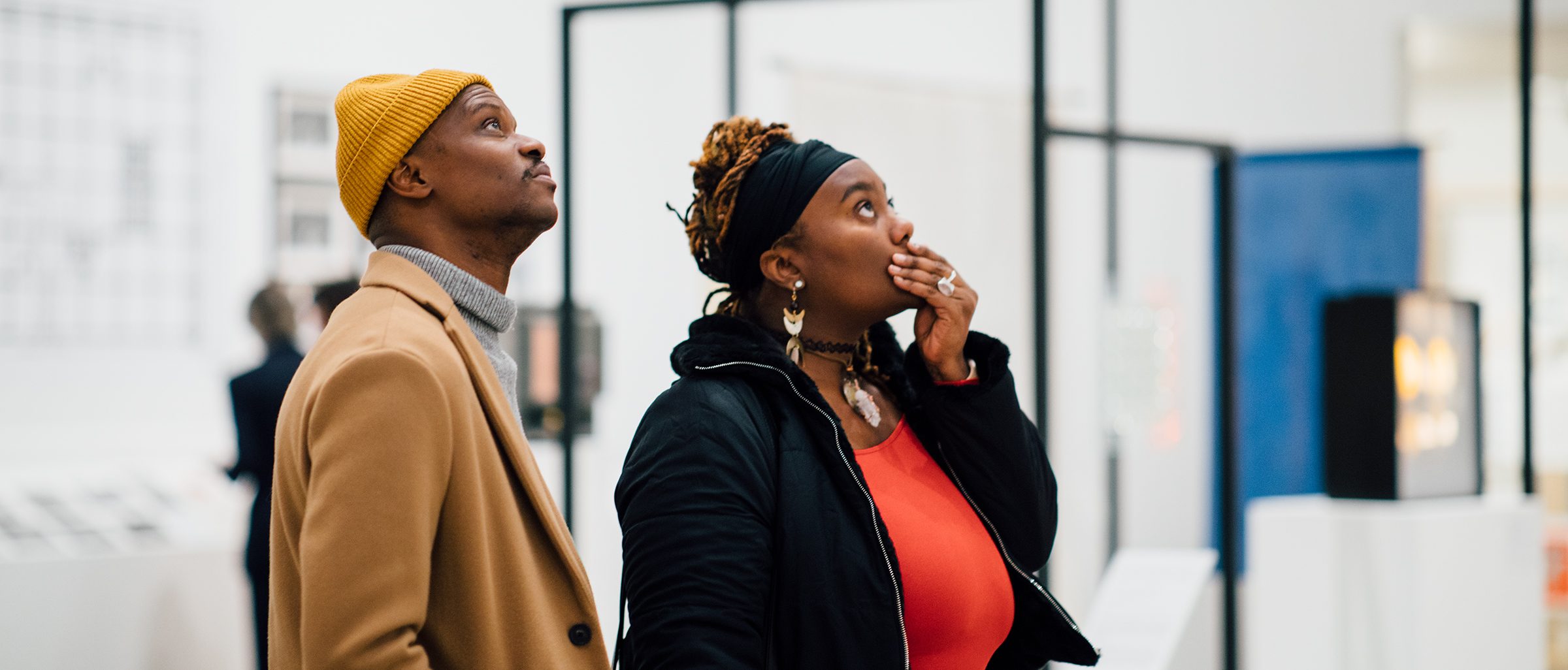 One adult wearing a camel colored beanie and coat and another adult wearing a headband and red top with a black coat, looking up at artwork in an exhibit at the Walker Art Center