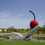 Large outdoor park sculpture of a cherry atop a spoon, floating over a small man-made lake, surrounded by grass, trees, and buildings