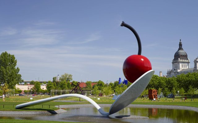 Large outdoor park sculpture of a cherry atop a spoon, floating over a small man-made lake, surrounded by grass, trees, and buildings