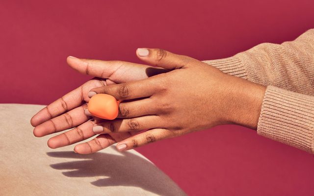 A pair of medium-skinned hands with tan colored sleeves, molding orange clay against a tan and red background