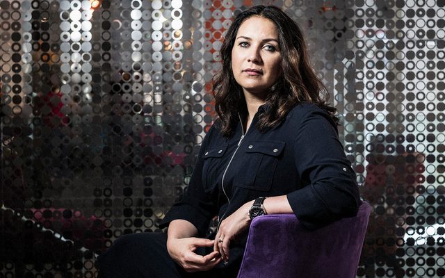 A long haired fair skinned adult sits in a purple chair with a silver background and turns back to camera