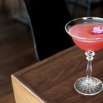 A long-stemmed glass drink with red fluid and a purple flower atop a brown table next to a black chair