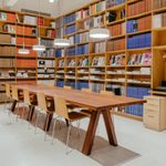 a long wood table with 10 chairs around it and 4 lights above it, in a room with large bookcase filled with green, brown, orange, blue, and white books