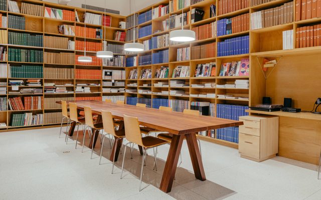 a long wood table with 10 chairs around it and 4 lights above it, in a room with large bookcase filled with green, brown, orange, blue, and white books