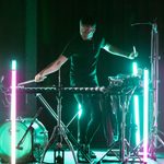 One person with light skin and short hair plays a xylophone with blue and pink vertical fluorescent lights standing next to them.