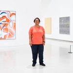 An adult with a coral shirt and dark pants, poses and smiles for a photo in front of displayed artwork in a museum