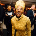 An adult with dark skin, a gold bodysuit, and turbin smiles at the viewer while surrounded by dancing people at a party.