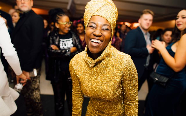 An adult with dark skin, a gold bodysuit, and turbin smiles at the viewer while surrounded by dancing people at a party.