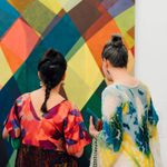 dev2024ag0921cl_086 Two people in colorful dresses look at a large painting displayed on a wall