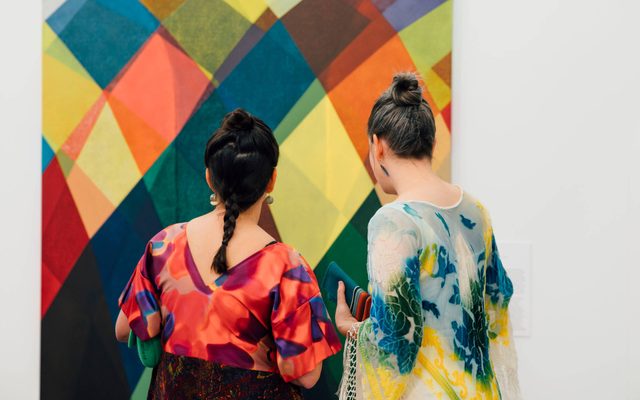 Two people in colorful dresses look at a large painting displayed on a wall