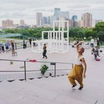 epp2019tftn0725_024 An adult with brown skin, wearing yellow pants and a flowy white top is mid-pose on stairs, a city view and sculpture and people in the background.