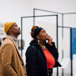 ex2023mr_opn_044 One adult wearing a camel colored beanie and coat and another adult wearing a headband and red top with a black coat, looking up at artwork in an exhibit at the Walker Art Center
