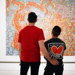 A taller adult has their hand on the lower back of another adult wearing a heart Keith Haring Tshirt, while looking at a displayed painting.