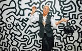 An adult poses in front of a black and whtie Keith Herring inspired wall, holding a leather skull bag, wearing daisy glasses and a fabulous outfit