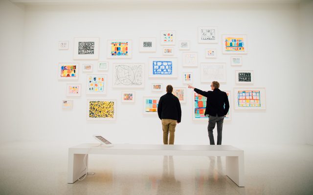 One adult points at a collection of paintings on a wall to another adult