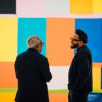 Two adults are talking to each other in front of a painting on the wall
