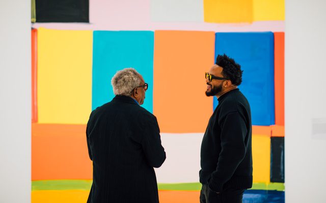 Two adults are talking to each other in front of a painting on the wall