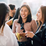 ex2024wp_opn_042 Three young women smile and chat together at an indoor social gathering or event.